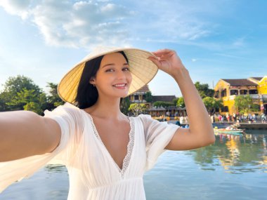 Beyaz elbiseli ve şapkalı güzel Asyalı kadın Hoi An, Vietnam 'da nehrin kenarında selfie çekiyor. Yüksek kaliteli seyahat, turizm, yaşam tarzı ve afiş veya tanıtım içeriği.