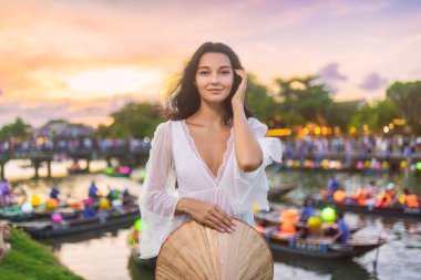 Hoi An 'da günbatımında fenerle aydınlatılmış teknelerin yanında geleneksel Vietnam konik şapkası tutan beyaz elbiseli genç bir kadın. Seyahat, moda veya kültür turizmi kampanyaları için ideal.