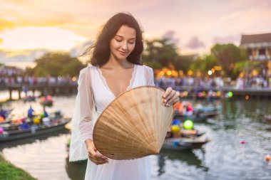 Gün batımında Hoi An nehrinde fener teknelerinin yanında geleneksel Vietnamlı konik şapkasına bakan akıcı beyaz elbiseli güzel bir kadın. Moda ve kültür turizmi için ideal.