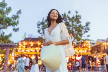 Beyaz elbiseli, elinde Vietnamlı koni şapkalı gülümseyen bir kadının fener gece piyasa portresi. Arkasında parlak ışık ve kalabalık var. Seyahat reklamları ve online yaşam tarzı kullanımı için ideal..