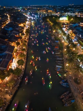 Hoi An 'ın hava gece görüşü Thu Bon Nehri' nde sürüklenen fener teknelerini, parlak yansımalarını ve nehir kenarındaki caddeleri gösteriyor. Yüksek kaliteli görüntü, seyahat reklamları, turizm ve festival temaları için ideal.