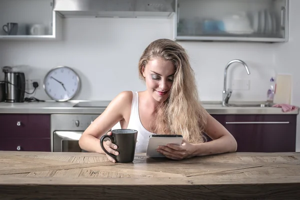 Блондинка сидит за кухонным столом, используя планшет и держа кружку в правой руке — стоковое фото