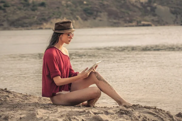 Młoda kobieta siedzi na plaży, czytając coś na tabletki — Zdjęcie stockowe