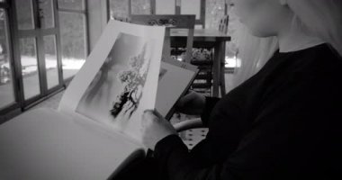 An attractive Moroccan Arab woman enjoys a photography art coffee-table book, turning pages. Art education. Leisure activity, enjoying free time at home. Black and white video.