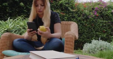 Woman surfs the web, sends text message on her phone while eating an apple, enjoying leisure time at home in the garden. Real-time footage.
