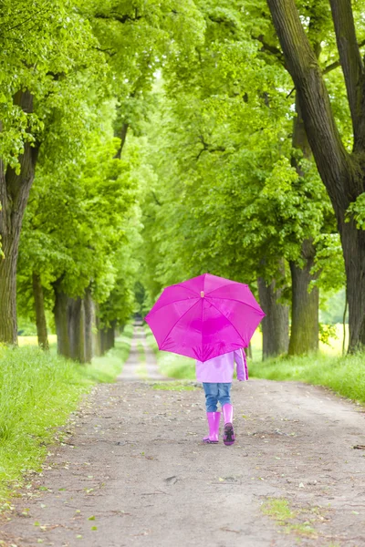 Little girl wearing rubber boots with umbrella in spring alley — Stock Photo, Image