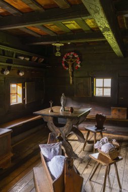 Ahşap mobilyalar, dini semboller ve pencereden gelen güneş ışığı olan köy odası.