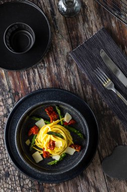 Bir tabak spagetti, yeşil kuşkonmaz, kurutulmuş domates ve ahşap masada parmesan talaşı.