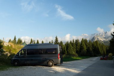 Çam ağaçları ve dağlarla çevrili çakıllı bir park yerine park etmiş bir karavan.