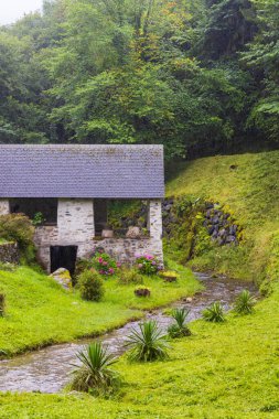 Tarihsel taş bina, yemyeşil bir doğal ortamda akan bir derenin yanında duruyor.