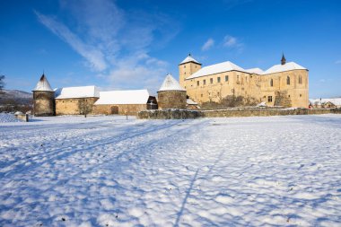 Taş duvarları ve parlak mavi gökyüzünün altında karla kaplı kuleleri olan Kapusany Kalesi