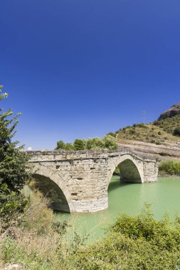 Açık mavi gökyüzünün altında yeşil suyun üzerinde birden fazla kemer bulunan antik taş köprü.