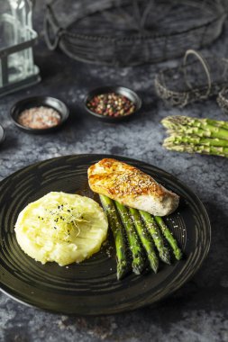 Tavuk göğsü, patates püresi ve siyah tabakta kuşkonmaz. Sağlıklı ve gurme bir akşam yemeği sunuyor.