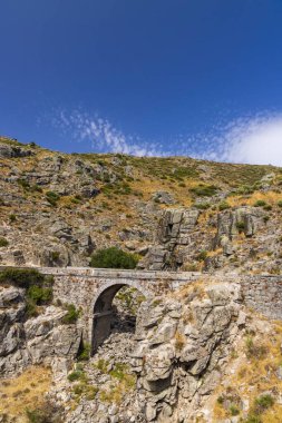 Tarihsel taş kemer köprüsü berrak mavi gökyüzünün altında kayalık, kurak bir dağ manzarasına inşa edildi.