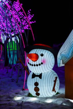 Kardan adam neşeli ışıklar ve tatil ruhuyla karlı bir akşamı aydınlatıyor.