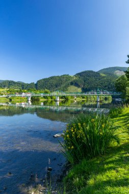 Yansımaları olan nehir boyunca uzanan truss köprüsü, yemyeşil dağlar ve açık mavi gökyüzü.