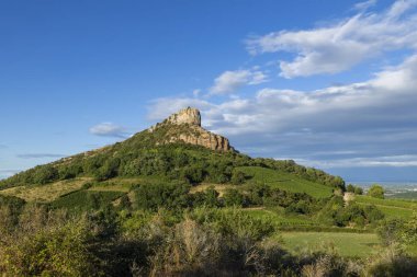 Solutre Kayası, saran üzüm bağlarının ve mavi gökyüzünün altındaki yeşil tepelerin üzerinde duruyor.