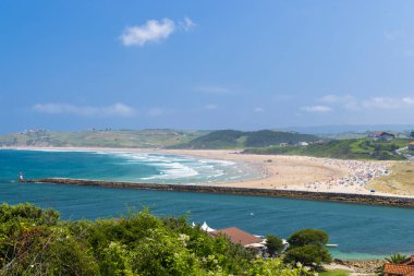Açık bir gökyüzünün altında dalgalı su ve deniz feneri olan kalabalık bir sahil ve körfez.