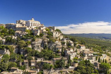 Gordes köyü binaları mavi gökyüzünün altındaki yeşil manzarayla çevrili kayalık bir yamaçta tırmanıyor.