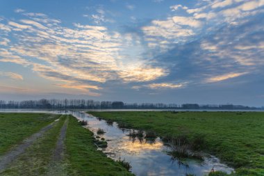 Yeşil alanlar, kirli bir patika ve Fransa 'nın Sougeal kentindeki bulutlu günbatımı gökyüzünü yansıtan bir su kanalı.