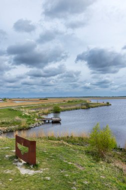 Kırsal alan, ahşap bir iskelesi olan bir gölü, çevreleyen tarlaları ve bulutlu gökyüzünü gösteriyor.