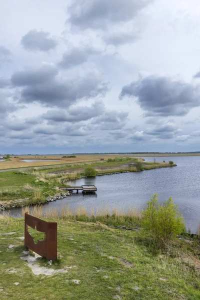 Kırsal alan, ahşap bir iskelesi olan bir gölü, çevreleyen tarlaları ve bulutlu gökyüzünü gösteriyor.