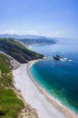 Engebeli bir kıyı şeridi, çakıl taşı plajı ve canlı mavi okyanus sularının manzarası.
