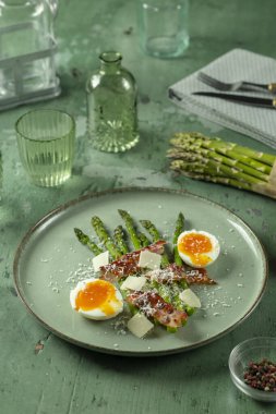 Çıtır pastırmalı taze kuşkonmaz, kaynamış yumurta ve yeşil tabakta rendelenmiş parmesan peyniri.