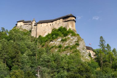 Orava Kalesi, açık bir gökyüzünün altındaki yemyeşil bir ormana bakan yüksek bir kayanın üzerine inşa edilmiş bir ortaçağ kalesi.