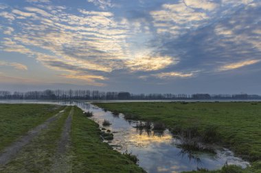 Çamur yolu ve dar kanallar sel basmış alanlarda dramatik bir gündoğumu gökyüzü altında dönüyor.