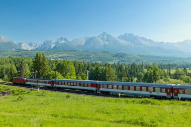 Kırmızı ve beyaz tren, mavi gökyüzünün altında yeşil tepeler ve dağlarla tren raylarında ilerliyor.