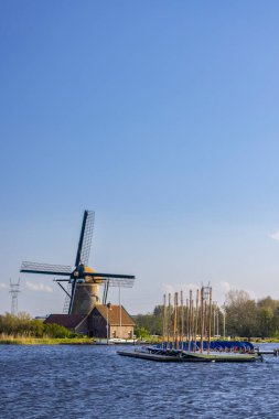 Klasik yel değirmeni, açık mavi gökyüzünün altında rıhtıma yanaşmış küçük teknelerle suyun kenarında duruyor.