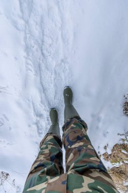Derin kış karlarında yürüyen, kamuflaj pantolonları ve lastik botlar giyen bir adam.
