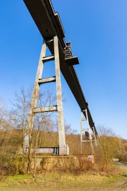 Açık mavi gökyüzünün altında ağaçlık bir vadiyi kaplayan yüksek beton yapı.