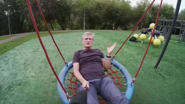 Seorang pria berayun di taman bermain dan berbicara di kamera — Stok Video