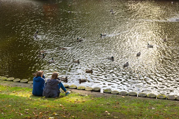 Duas meninas descansando no banco de uma lagoa com patos — Fotografia de Stock