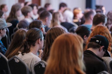 Katılımcılar konferans odasında sıra sıra otururlar, sahneye, çeşitli gruplara, günlük kıyafetlere, meşgul seyircilere, konuşma ve sunumlar sırasında konsantrasyon ve sessizlik anlarına odaklanırlar.