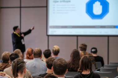 Sunucu konferans sırasında ilgili seyircilere hitap ederken, büyük ekran slaytları ve mavi kalkan simgesini gösteriyor