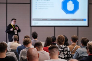 Katılımcılar dikkatle dinlerken konuşmacı odanın önünde sunar. Geniş ekran, çeşitli katılımcılarla mavi seminer ayarlarını gösteriyor