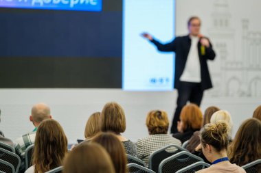 Sahnedeki bulanık konuşmacı modern konferans odasında oturan kalabalığa sunum yapıyor. Büyük ekranda profesyonel öğrenme atmosferi yaratan görseller gösterilirken dikkatli katılımcılar dinlerler