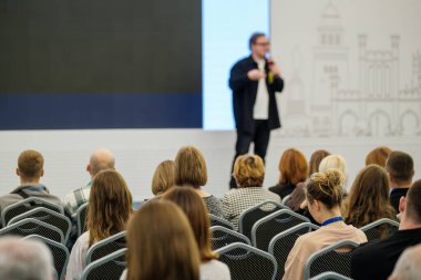 Konferans salonunda kalabalık dikkatle dinlerken sahnede konuşma yapan bir sunucu. Sıralar dolusu sandalye. Ufuk çizgisi. Profesyonel ruh hali öğrenme.