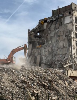 Yıkım sahnesi, turuncu kazıcının beton bina, moloz ve tozu yerle bir ettiğini gösteriyor. Havayı dolduruyor, açığa çıkan çelik çubuklar yıkımı ve ağır makineleri işbaşında gösteriyor..