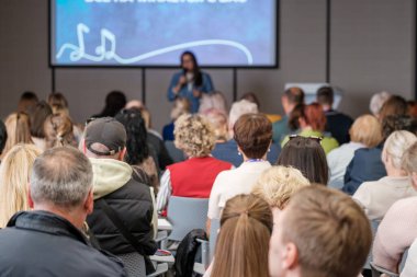 Kalabalık konferans odasında oturmuş arkada büyük ekran sunumuyla hoparlörleri dinliyor.