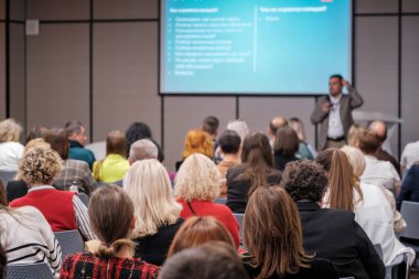 Profesyonel konferans kalabalığı slaytları gösteren büyük ekranlı sunucuyu dinliyor. Katılımcılar odaklanır, notlar alır ve parlak modern konferans salonunda fikirleri tartışırlar..