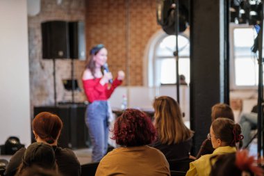 Kırmızı top giyen konuşmacı, tuğla duvar arka planında sıcak ışıklandırma ve katılımcı katılımcıları ile gündelik etkinlik sırasında dinleyicilere hitap ediyor