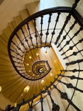 Yukarıdan gelen sarmal merdivenlerde koyu demir balyalar ve parlayan kolye ışıkları dramatik kıvrımlar ve sıcak bir ortam yaratıyor.