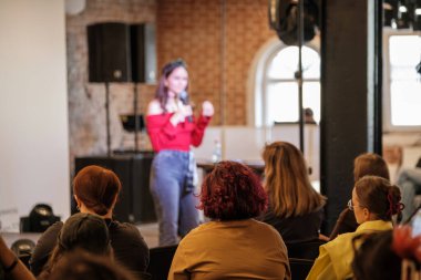 Kırmızı omuzlu sunucu, tuğla duvarlı salonda özenli seyircilerle konuşuyor. Sahne ekipmanları ve parlak pencereler dinamik öğrenme anını çerçevelerken seyirciler oturup dinledi