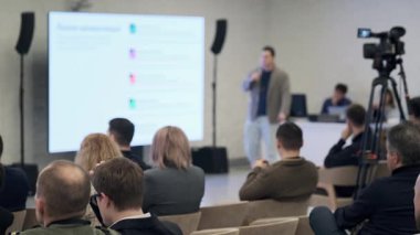 Konuşmacı, modern konferans odasında özenli dinleyicilere sunar. Kamera kayıtları olarak büyük ekran slaytları görüntüler.