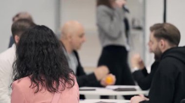 İnsanlar parlak ofis ortamında takım tartışması sırasında sohbet ederler. Ön planda pembe ceketli kadınlar görünür. Diğerleri ise gelişigüzel bir iş toplantısı gibi dinler ve plan yapar.