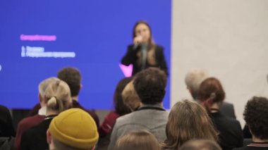 Sahnedeki bulanık konuşmacı, canlı mavi ekran ile kapalı konferansta kalabalığı meşgul ederek profesyonel atmosfer yaratıyor.